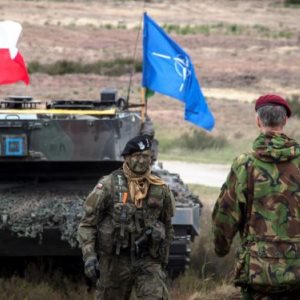 Ein Soldat aus Polen und ein Soldat aus den Niederlanden stehen sich vor einem Panzer, auf dem die Flaggen Polens und der NATO gehisst sind, gegenüber.