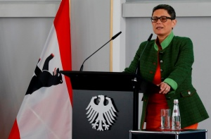 Eine Frau spricht an einem Rednerpult, das ein Bundesadler ziert. Im Hintergrund ist die deutsche Fahne Berlins erkennbar.