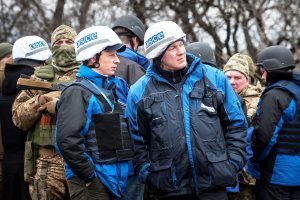 Das Foto zeigt mehrere uniformierte Angehörige der OSZE-Beobachtermission in der Ukraine und mehrere bewaffnete und zum Teil vermummte Soldaten. Zwei der Beobachter schauen sich um, während ihnen ein Vermummter über die Schulter schaut.