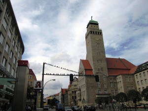 Rechts im Bild ragt das steingemauerte Rathaus Neukölln mit seinem Turm auf, links die Wand eines mehrstöckigen modernen Gebäudes; in der Mitte ist eine breite Straße erahnbar und eine Verkehrsampel, auf der zahlreiche Tauben sitzen, sichtbar.