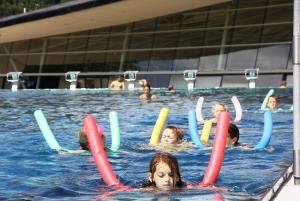 Mehrere Kinder schwimmen mit langen dünnen zylindrischen bunten Schwimmhilfen in einem Schwimmbecken; im Hintergrund ragt hinter Schwimmstartblöcken die Glasfassade des Schwimmbads auf.