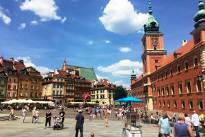 Renovierte mehrstöckige Altbauten und ein Klinker-Prachtbau begrenzen einen sommerlich anmutenden Platz mit Menschen darauf, überspannt von einem blauen Himmel mit vereinzelten Kumuluswolken.