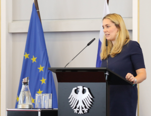 Victoria Wieninger steht an einem Pult mit dem Bundesadler darauf und spricht in ein Mikrofon; im Hintergrund stehen die Flaggen der EU und Finnlands.