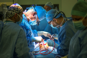 Mehrere mit Operationskleidung und Mundschutz verhüllte Ärzte beugen sich mit Operationswerkzeug über einen verdeckt liegenden Patienten.