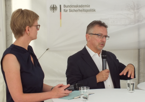 Die Journalistin Ulrike Scheffer und der Präsident der BAKS Dr. Karl-Heinz Kamp stehen nebeneinander an einem Stehtisch; im Hintergrund hängt ein Banner der BAKS, und Kamp spricht in ein Mikrofon in seiner Hand.