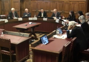 In einem Ausschusssaal des britischen Parlaments befinden sich Ausschussmitglieder in einer Videokonferenz mit BAKS-Präsident Karl-Heinz Kamp, dessen Bild auf mehreren Monitoren übertragen wird.