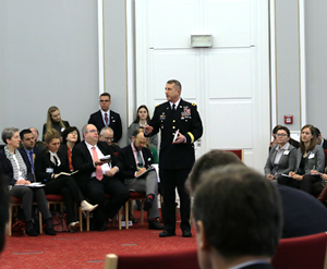 A military officer is standing among an audience of several both uniformed and business-dressed people and speaks into a microphone.
