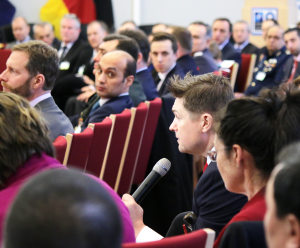 Sitting among other people within a row of chairs, a business-dresses young man talks into a microphone while several neighbors look at him.