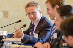 Ein Mann im Anzug beugt sich über einen Konferenztisch mit Mikrofonen zu Gesprächspartnern hinüber und spricht gestikulierend.