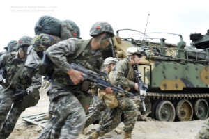 In Tarnanzügen gekleidete Soldaten verschiedener Staaten mit Kampfausrüstung rennen an einem Panzerfahrzeug vorbei.