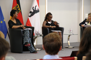 Drei Frauen sitzen auf einem Podium und sprechen miteinander vor einem Publikum. 