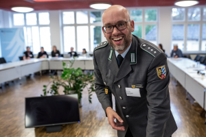 Ein lachender Mann in Uniform beugt sich über einen Tisch; im Hintergrund sitzen Menschen um ein Tischkarree.