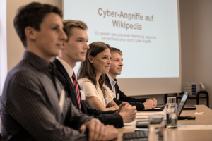 Vier junge Menschen sitzen an einem Tisch; im Hintergrund wird eine Präsentation mit dem Titel "Cyberangriffe auf Wikipedia"  auf eine Leinwand projiziert.