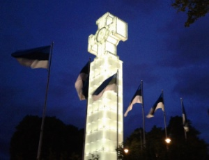 Eine Kreuzskulptur vor Nachtthimmel, dahinter wehen mehrere Flaggen im Wind.