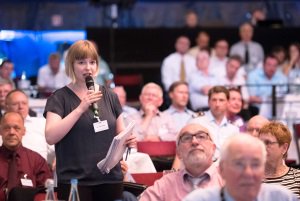 Eine junge Frau mit Unterlagen in der Hand spricht einen Diskussionsbeitrag in ein Mikrofon.