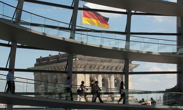 In der Kuppel des Reichstagsgebäudes laufen Menschen die spiralförmige Rampe entlang; außerhalb ist einer der Ecktürme mit einer wehenden Deutschlandflagge erkennbar.