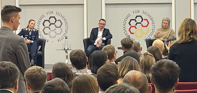 Eine Soldatin und zwei Zivilpersonen sitzen auf einem Podium und blicken zu einem Publikum, aus dem ein Mann und eine Frau stehend mit ihnen interagieren.