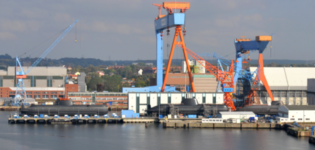 Auf einem Werftgelände am Wasser liegen mehrere Unterseeboote im Bau an Land; darüber ragen große Werftkräne auf.