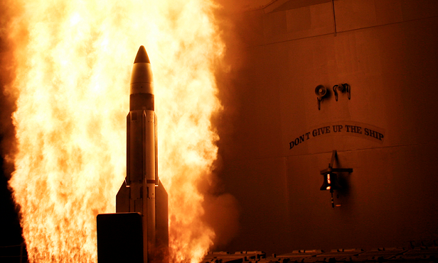 Eine Rakete verlässt vor einem Feuerschweif einen Startkanister eines Kriegsschiffs; daneben ist eine Schiffsglocke mit dem Motto des Schiffs erkennbar.