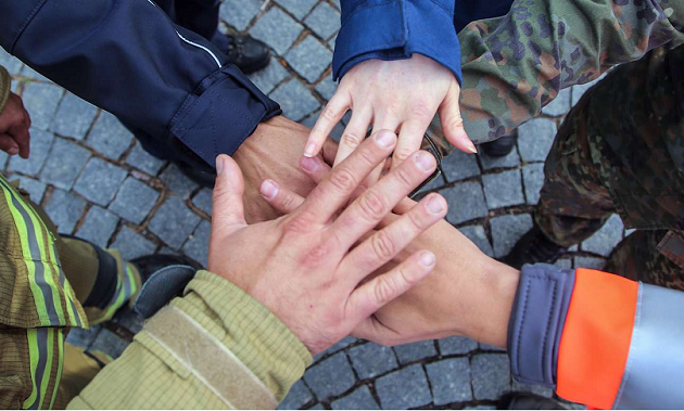 Fünf Personen, die verschiedene Uniformen tragen, stehen im Kreis und halten ihre Hände übereinander.