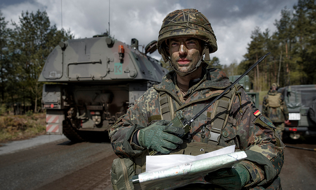 Ein Bundeswehrsoldat in Kampfausrüstung steht hinter dem Heck einer Panzerhaubitze 2000; in den Händen hält er eine Karte und ein Funkgerät.