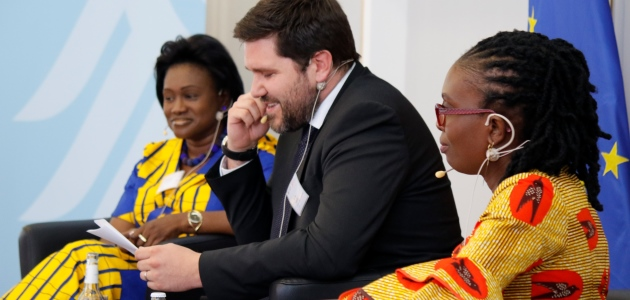 Zwei traditionell afrikanisch gekleidete Frauen und ein geschäftlich gekleideter Mann, jeweils mit Ohrbügelmikrofonen, sitzen gemeinsam auf einem Podium; links ragt eine Bundesadlerschwinge ins Bild, rechts die Europaflagge.