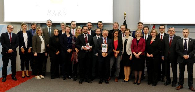 Zahlreiche junge und geschäftliche gekleidete Menschen stehen auf einem Podium und blicken zum Betrachter.