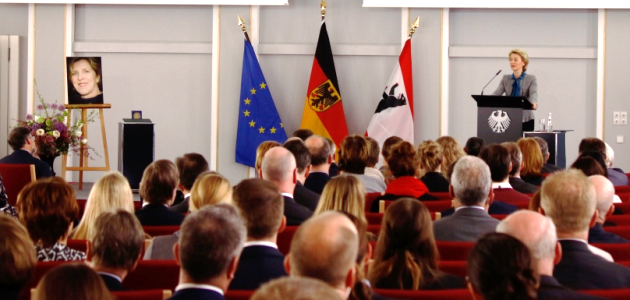 Im Historischen Saal der BAKS steht links eine Staffelei mit dem Porträt Sylke Tempels sowie einem Blumenstrauß. Rechts im Bild steht Verteidigungsministerin von der Leyen an einem Rednerpult.