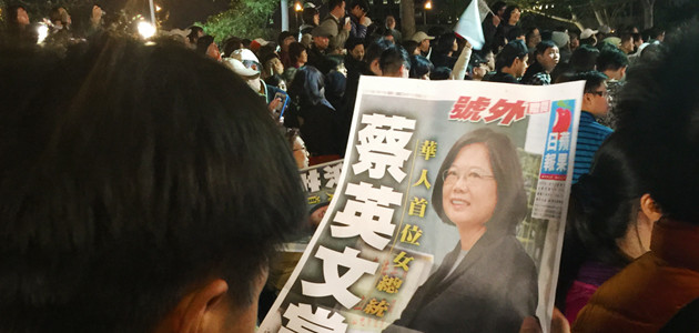 Ein Mann blickt am Tag der taiwanesischen Wahl auf eine Zeitung, die Tsai Ing-wen abbildet, die Spitzenkandidatin der DPP bei den Präsidentschaftswahlen. Im Hintergrund stehen Menschen mit Fahnen.