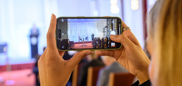 Ein von zwei Händen horizontal gehaltenes Smartphone zeigt ein Kamerabild aus dem Historischen Saal der BAKS, in dem zwei schemenhaft erkennbare Personen vor Publikum sprechen.