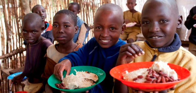 Eine Gruppe afrikanischer Kinder sitzt mit gefüllten Tellern in den Händen in einem Gebäude aus weidengeflechtartigen Holzwänden und blickt zum Betrachter.