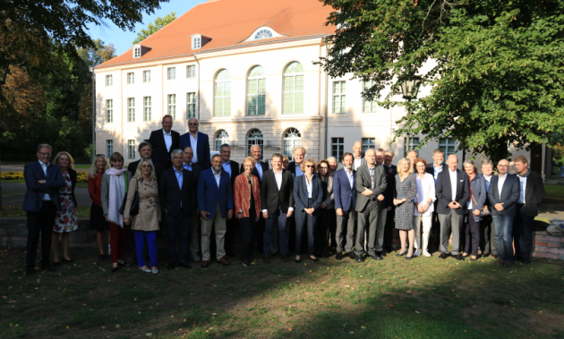 Eine Gruppe geschäftlich gekleideter Menschen steht auf dem Rasen zwischen Bäumen vor einem Schloss.