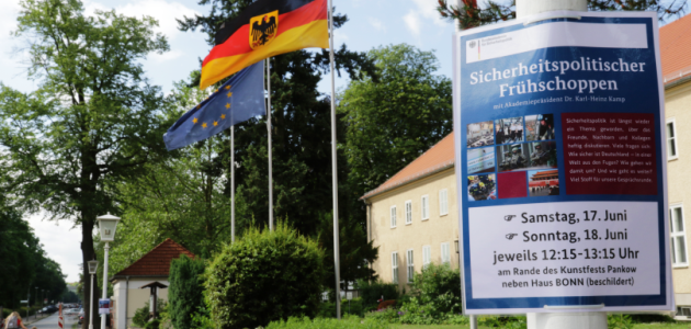 Vor dem Haus Berlin der BAKS wehen die Flaggen der EU, Deutschlands und Berlins; an einem Laternenmast recht hängt ein Plakat mit der Aufschrift "Sicherheitspolitischer Frühschoppen".