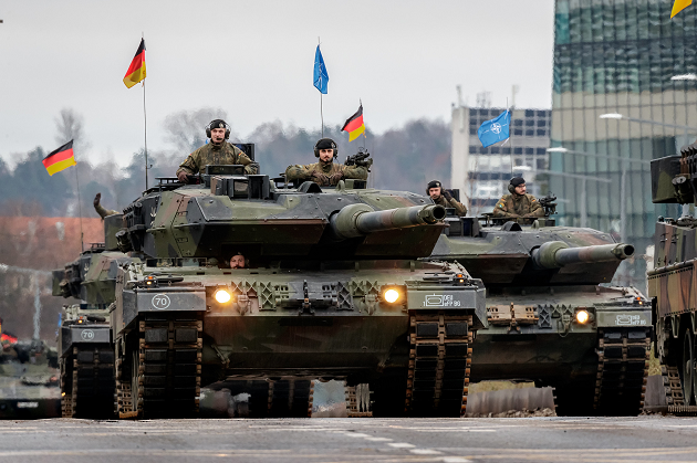 Bundeswehr-Kampfpanzer Leopard 2 mit Flaggen Deutschlands und der NATO fahren in Paradeformation auf einer Straße in Litauen anlässlich des 105. Geburtstags der Streitkräfte Litauens.