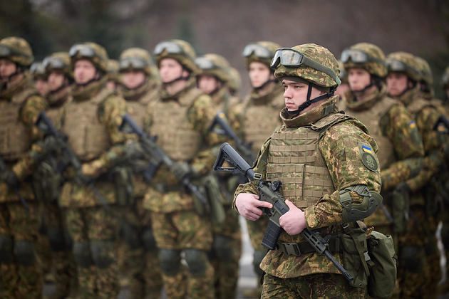 Ukrainische Soldatinnen und Soldaten bewaffnet und in Kampfbekleidung stehen angetreten in einer Reihe; ein Soldat steht im Vordergrund.