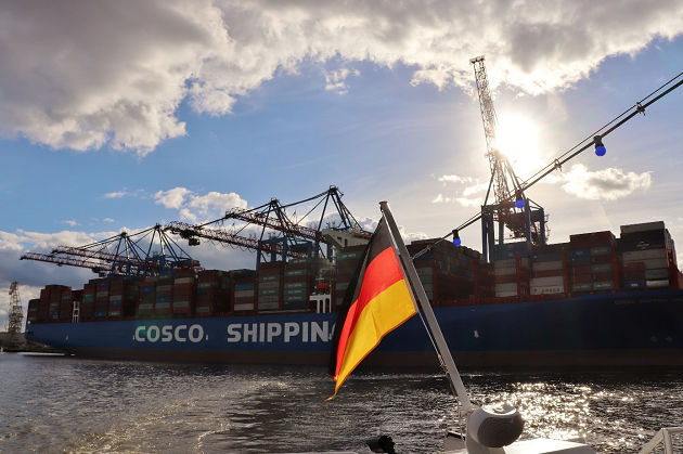 Vor einem großen Containerschiff mit der Aufschrift COSCO ist die Flagge Deutschlands am Heck eines anderen, kleineren Wasserfahrzeugs erkennbar.