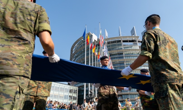 Mehrere Soldaten halten eine ausgebreitete EU-Flagge; im Hintergrund ragt das Plenargebäude des EU-Parlaments auf.