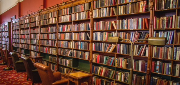 Von links hinten nach rechts vorn füllt ein nahezu wandhohes altes Bibliotheksregal voller Bücher das Bild aus; davor stehen Sessel.