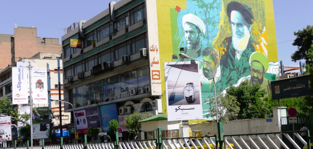 Das Foto zeigt, von einer Straße aus betrachtet, die Seitenwand eines Gebäudes, die ein großes Wandbild trägt, welches Ajatollah Chomeini und weitere bekannte Personen Irans zeigt; davor steht ein Werbeplakat für einen japanischen Uhrenhersteller.