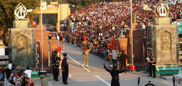 Beiderseits eines Grenztores sind verschieden gekleidete Soldaten aufmarschiert und halten ein Zeremoniell ab. Die Szenerie wird durch hunderte Zuschauer auf einer Tribüne verfolgt.
