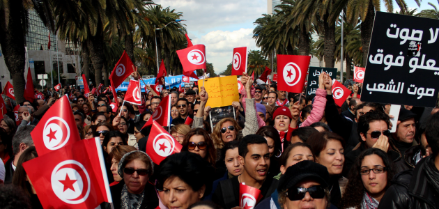 Auf einer Straße gesäumt von Palmen steht eine große Menschenmenge, die zahlreiche Flaggen Tunesiens und Plakate in arabischer Sprache mit sich führt.