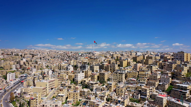 Das Bild zeigt ein Panorama der Stadt Amman mit einer Flagge Jordaniens im Mittelpunkt.