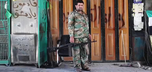 Ein Kämpfer der \"Freien Syrischen Armee", bewaffnet mit einem Maschinengewehr, vor einer verschlossenen, mit Graffiti besprühten, Ladenfront.