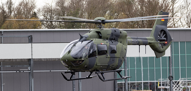 Ein Hubschrauber vom Typ Airbus H145M in Tarnlackierung der Bundeswehr fliegt niedrig vor einem Hangargebäude.