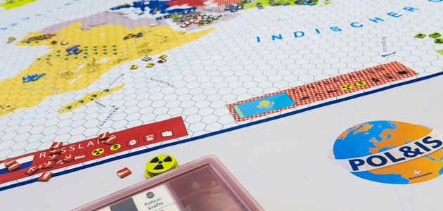 Das Bild zeigt die Welt Pol&IS als Spielbrett. Daneben liegen Spielkarten und Spiel-Chips mit aufgedrucktem Atomzeichen, rechts davon ist die Aufschrift Pol&IS zu lesen.