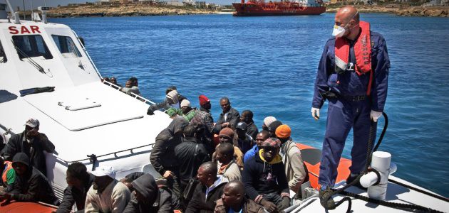 Flüchtlinge aus Afrika sitzen in einem Boot der italienischen Küstenwache; ein Angehöriger der Küstenwache steht am Bug und vertäut das Boot.