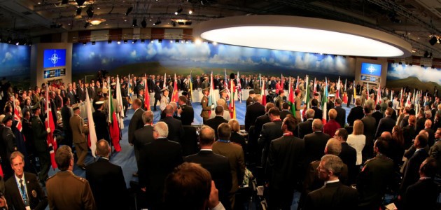Zahlreiche Menschen stehen in einem zum Teil abgedunkelten Konferenzsaal mit Projektionen der NATO-Flagge an den Wänden.