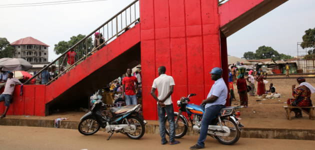 Zahlreiche afrikanische Frauen und Männder stehen und sitzen bei einem nach oben rechts aus dem Bild ragenden rot gestrichenen Treppenaufgang aus Beton. Vorn stehen zwei Krafträder; im Hintergrund ragt links ein Neubau, rechts improvisierte Stände auf.