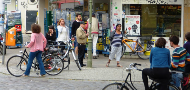 Vor einem Eckladengeschäft mit einer alten Straßenlaterne und Plasterstraße davor stehen und gehen zahlreiche Menschen verschiedener Herkunft und verschiedenen Alters, mehrere mit Fahrrädern; die gesamte Szene mutet durch Graffiti rauh und bunt an.