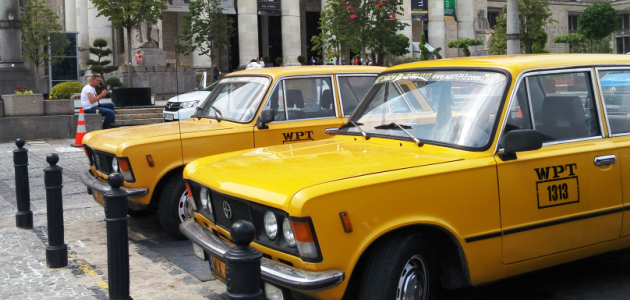 Im Vordergrund stehen zwei gelbe Taxis osteuropäischer Bauart; im Hintergrund ragt eine Säulenkollonade mit Plakaten zwischen den Säulen auf.